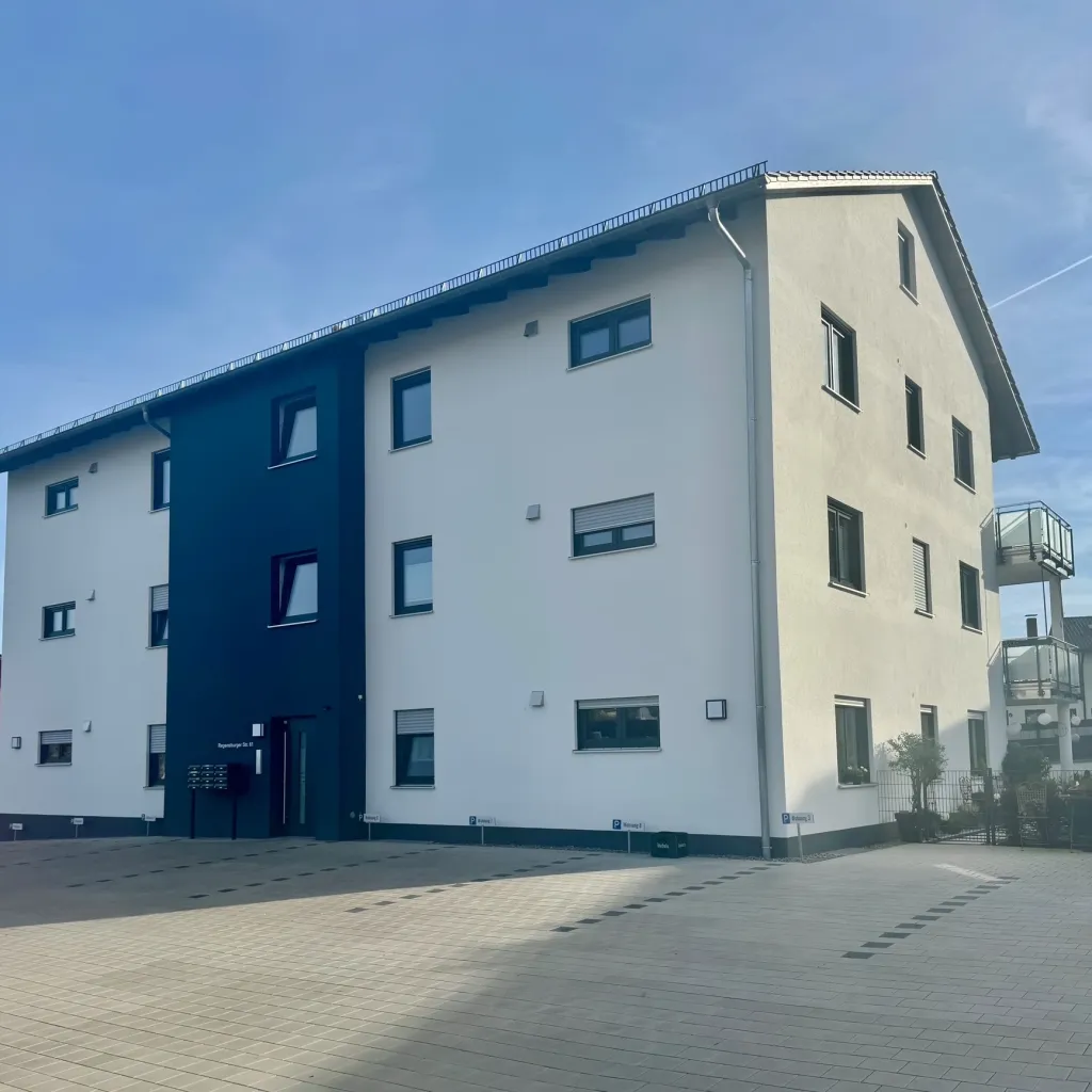 Weißes Mehrfamilienhaus mit dunkelblauem Treppenhaus und gepflastertem Vorplatz