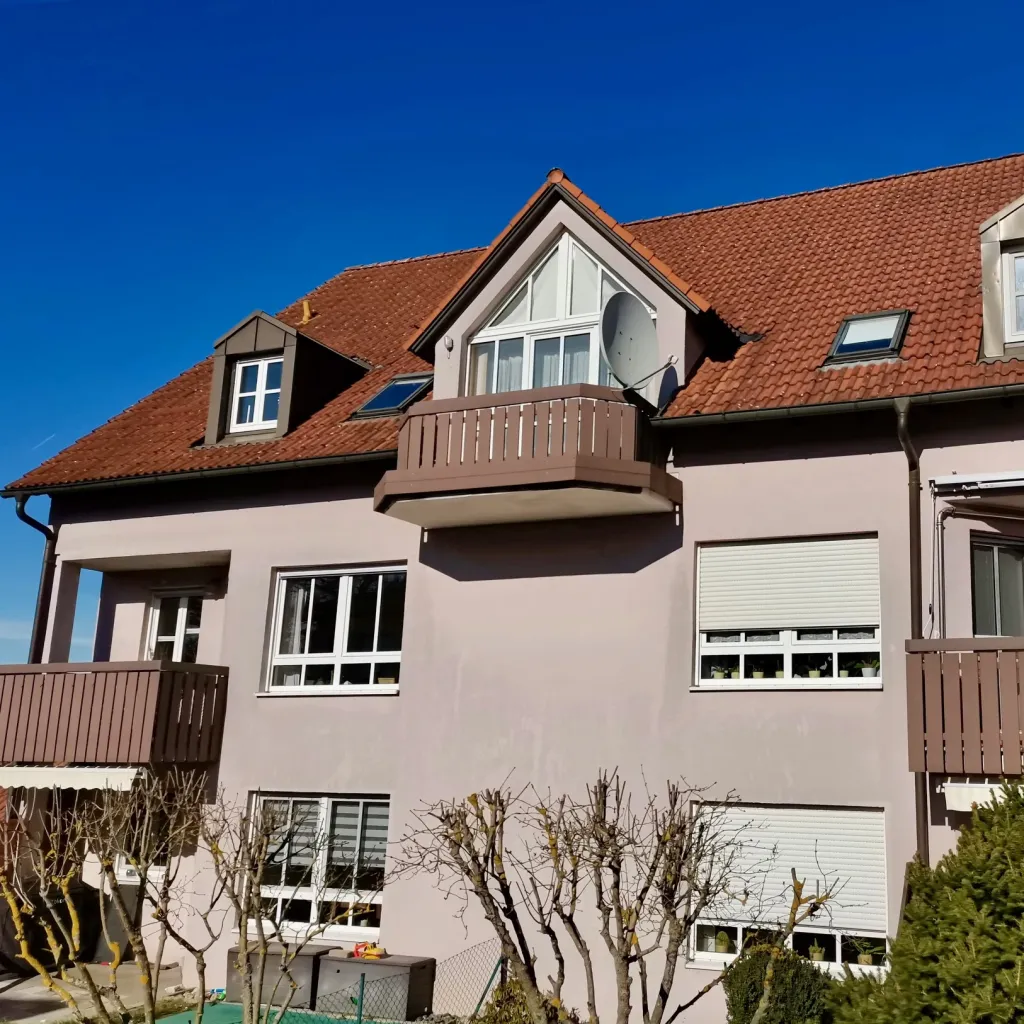 Altrosa Mehrfamilienhaus mit Holzbalkonen und rotem Ziegeldach vor blauem Himmel